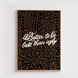Puede incluir: Un cuadro rectangular con marco que presenta un estampado animal de leopardo en marrón y negro. Centrado en la impresión está el texto cursivo blanco que dice, Better to be late than ugly. El marco es de color madera clara con un borde fino.