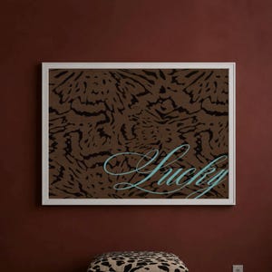 Peut inclure: Une œuvre d'art murale rectangulaire dans un cadre blanc présentant un motif animalier abstrait marron et noir avec le mot Lucky écrit en écriture cursive bleu clair sur la partie inférieure, placée au-dessus d'un pouf à motif léopard.