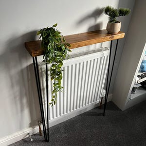May include: A wooden console table with a stained finish and black hairpin legs. The table is adorned with a trailing green plant and a small potted evergreen. The table is placed in front of a white radiator.