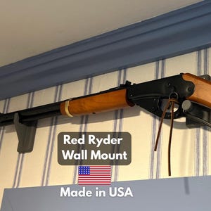 May include: A Red Ryder BB gun mounted on a wall. The gun has a wooden stock and a black metal barrel. The text "Red Ryder" is engraved on the stock. The wall mount is gray.
