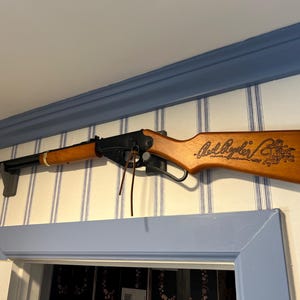 May include: A toy rifle in wood and black, mounted on a wall. The rifle features a light brown wooden stock and barrel, with the words "Red Ryder" etched into the stock. It is mounted on a grey bracket.