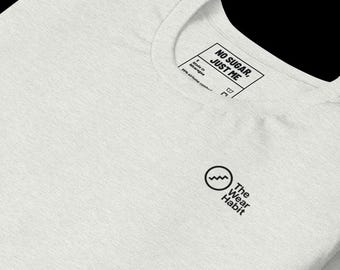 May include: A light gray t-shirt with a crew neck. The shirt features a small black circular logo with a wave design and the text "The Wear Habit". The tag reads "NO SUGAR, JUST ME".