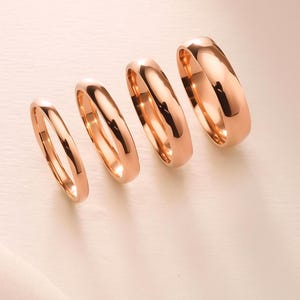 May include: Four rose gold wedding bands of varying widths are arranged diagonally. The rings have a smooth, polished finish, reflecting light. The bands are displayed on a light pink surface, creating a soft, elegant aesthetic.