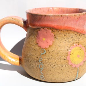 May include: Handmade ceramic mug with a speckled tan body, a pink rim, and a matching handle. The mug is decorated with pink and yellow flower designs and the word "peace" in a teal script.