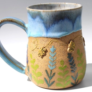 May include: A ceramic mug with a speckled tan body featuring painted green and blue floral stems and raised yellow flower accents. The upper portion of the mug and the handle are glazed in a smooth, glossy blue that drips down onto the tan base.