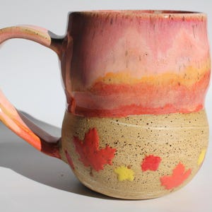 May include: A ceramic mug with a rounded base and a large handle. The upper portion features a smooth, glossy pink and orange gradient glaze, while the lower portion has a speckled beige finish decorated with small, raised red and yellow maple leaves.
