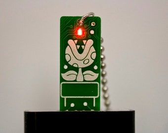 May include: Green circuit board keychain featuring a white Piranha Plant design. The keychain has a red LED light and a silver ball chain. The design is reminiscent of a classic video game.