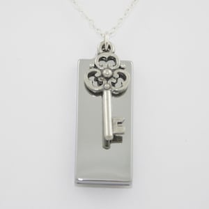 May include: Silver-toned key pendant necklace. The pendant features a rectangular plate with a decorative key charm. The key has an ornate top and a detailed key bit. The necklace chain is also silver.