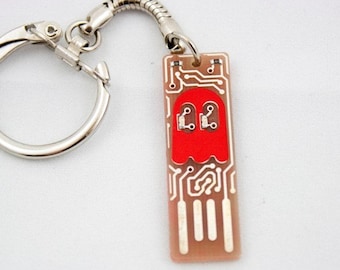 May include: A novelty keychain featuring a rectangular pendant with a circuit board design. A red Pac-Man ghost is in the center. The keychain has a silver-colored key ring and a flexible metal chain.