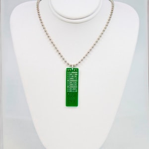 Może przedstawiać: Srebrny naszyjnik z łańcuszkiem z kulek i prostokątną, zieloną zawieszką. Zawieszka zawiera biały kod binarny. Naszyjnik jest prezentowany na białym popiersiu.