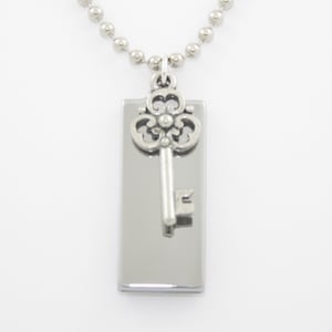 May include: A silver-toned necklace featuring a rectangular pendant with a decorative key charm. The key is positioned over the pendant, and the necklace has a ball chain. The jewelry is displayed against a white background.