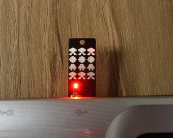May include: A black USB flash drive with a retro arcade game design featuring white pixelated alien figures. The drive has a small hole at the top and a red light is illuminated at the bottom. The background is a wooden surface.