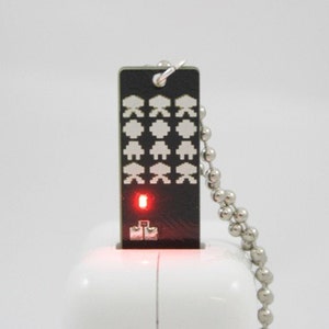May include: A black USB drive with a pixelated alien design and a silver chain. The drive has a red light illuminated at the bottom. The design features white pixelated aliens on a black background.