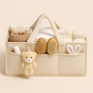 Könnte beinhalten: Cremefarbener Baby-Organizer-Korb mit weichem, strukturiertem Äußeren und flauschigem Griff. Der Korb enthält Babyutensilien wie Windeln, eine Strickmütze, Bürsten und einen Teddybären. Die Taschen enthalten eine Tube und Hasenohren.