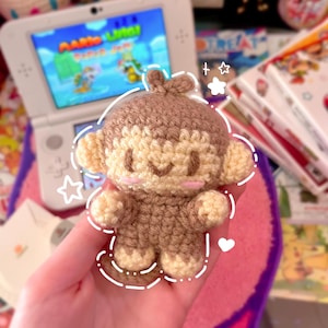 May include: A small, handmade crochet monkey plush toy featuring light brown and cream yarn. The monkey has a rounded head, small ears, and embroidered facial features with pink cheeks. It is decorated with white digital line art of stars and hearts.