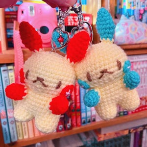 May include: Two handmade crochet keychains featuring yellow creatures with red ears and accents. One has blue accents, the other has red. Each keychain has a silver clasp and a heart-shaped charm with a star.