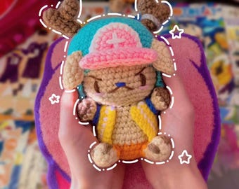 May include: A handmade crochet plush toy of a character with a blue hat, pink hat, and brown antlers. The toy has a yellow vest and orange shorts. The plush is held in two hands against a pink and purple background.