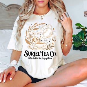 A woman wearing a white t-shirt with a graphic design and text, sitting on a bed with a potted plant in the background.