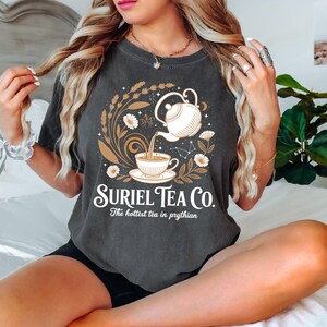A woman with long blonde hair is sitting on a bed, wearing a gray t-shirt with a graphic design of a teapot and flowers.