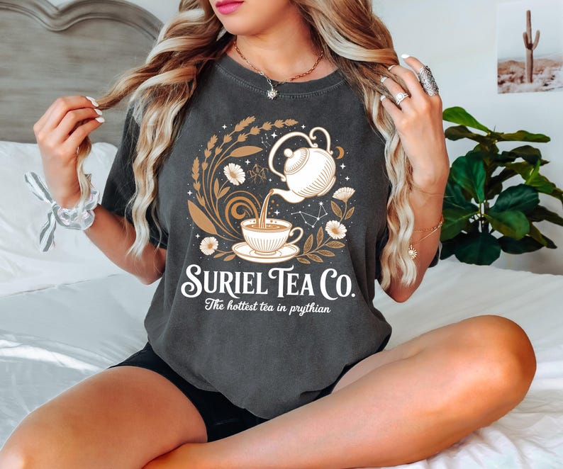 A woman with long blonde hair is sitting on a bed, wearing a gray t-shirt with a graphic design of a teapot and flowers.