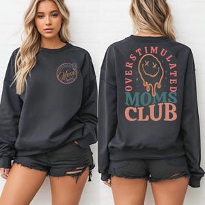 May include: A dark gray sweatshirt with the text "Moms Club" on the front and back. The front features a circular design with the words "Moms Club". The back has the words "Overstimulated Moms Club" with a melting smiley face graphic.