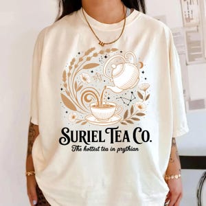 A person wearing a white t-shirt with a graphic design featuring a teapot and floral elements, along with the text "Suriel Tea Co. The hottest tea in Pyrhian".