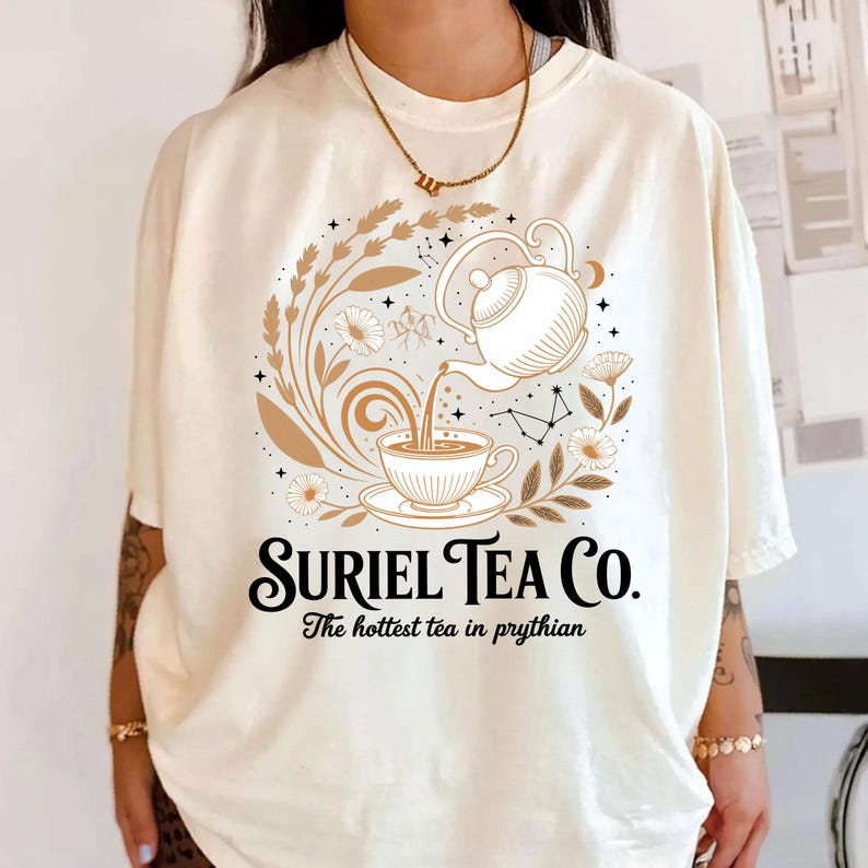 A person wearing a white t-shirt with a graphic design featuring a teapot and floral elements, along with the text "Suriel Tea Co. The hottest tea in Pyrhian".