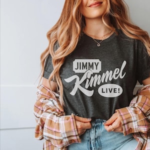May include: Dark gray t-shirt with the text "JIMMY Kimmel LIVE!" in white. The shirt is paired with a plaid button-down shirt in shades of brown, pink, and white. The person is wearing blue jeans and gold jewelry.