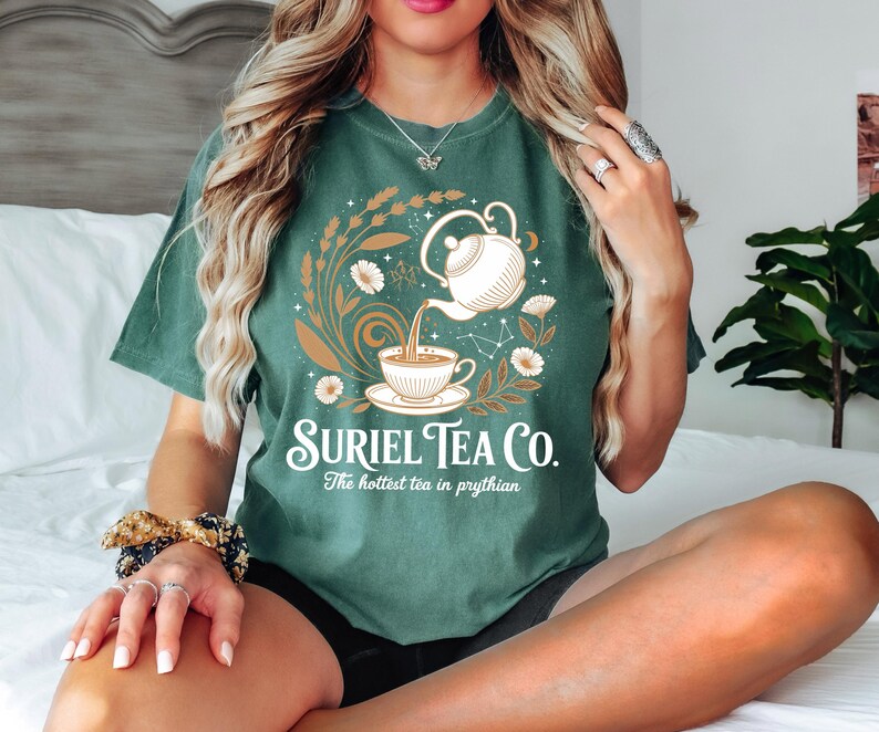 A woman wearing a green t-shirt with a graphic design of a teapot and flowers, sitting on a bed with a potted plant in the background.