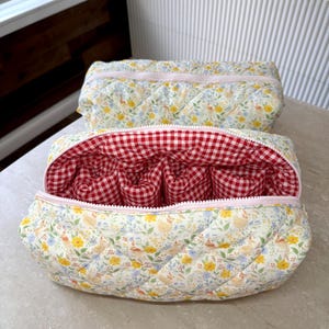 May include: Two quilted cosmetic bags. The exterior fabric features a floral and chicken pattern on a cream background. The interior of one bag is red and white gingham. Both bags have a pink zipper and are open to reveal compartments.