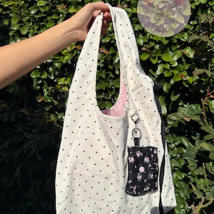 May include: A white tote bag with black polka dots, held up against a green leafy background. The bag has a pink gingham lining and a small black pouch with a floral pattern attached. A small keychain is attached to the bag.