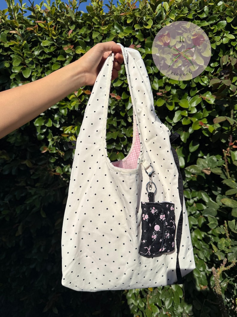 May include: A white tote bag with black polka dots, held up against a green leafy background. The bag has a pink gingham lining and a small black pouch with a floral pattern attached. A small keychain is attached to the bag.