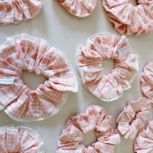 May include: A collection of pink scrunchies with various patterns. Some feature a gingham design, while others display a repeating pattern of small peaches. Several scrunchies have a white lace trim. Each scrunchie has a small white tag that reads "ricocoh".