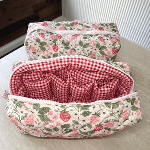 May include: Two quilted cosmetic bags. The exterior features a cream background with a strawberry and floral pattern. The interior of one bag is red and white gingham. Both bags have a white zipper.