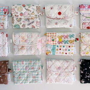 May include: A collection of quilted fabric pouches in various patterns and colors. Designs include floral, polka dots, and cartoon characters. Each pouch has a button closure and a small loop. The pouches are arranged in a grid.