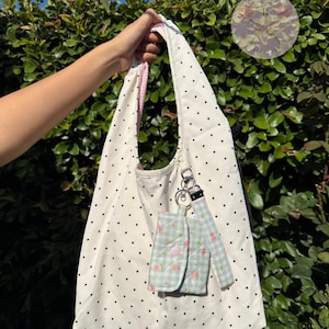 May include: A white tote bag with black polka dots, held by a hand. The bag has a pink gingham lining and a matching small pouch with a floral pattern and a pink button closure. A silver keychain is attached.