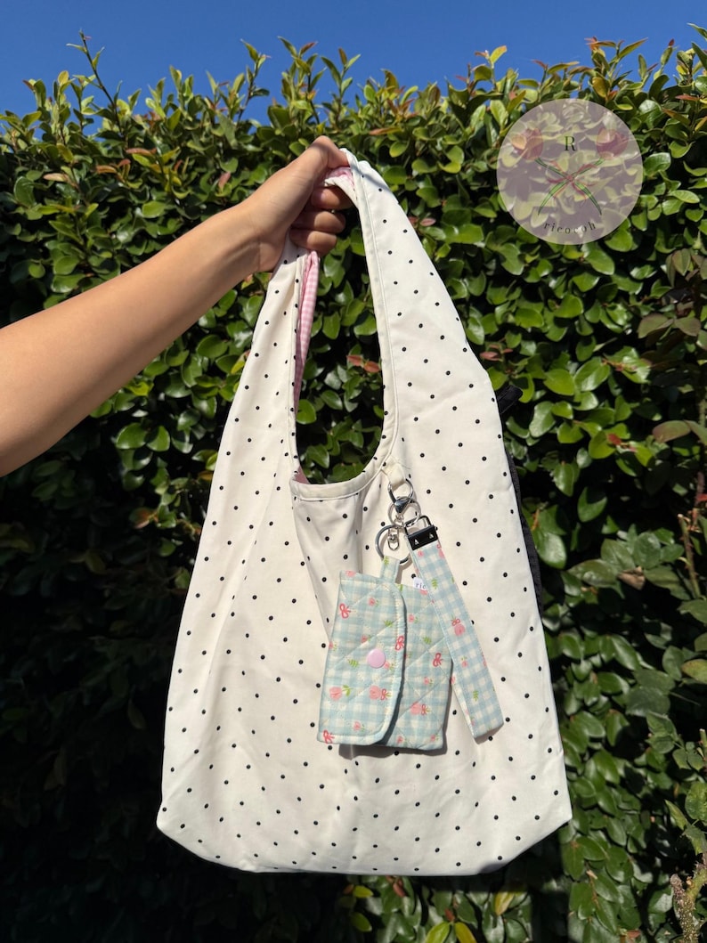 May include: A white tote bag with black polka dots, held by a hand. The bag has a pink gingham lining and a matching small pouch with a floral pattern and a pink button closure. A silver keychain is attached.