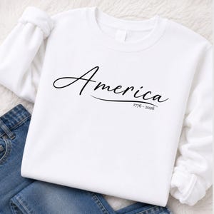 May include: A white long-sleeved sweatshirt featuring the word America in black cursive script across the chest, with the text 1776 - 2026 printed in a smaller black font underneath the word, paired with blue denim jeans.
