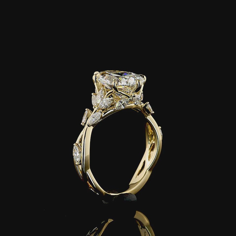 Puede incluir: Un anillo de compromiso de oro con un diamante grande y claro de talla ovalada como pieza central. El anillo presenta diamantes m&aacute;s peque&ntilde;os y detalles en forma de hojas a lo largo de la banda. El anillo est&aacute; sobre un fondo negro.