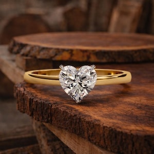 May include: A gold ring featuring a heart-shaped diamond. The clear, faceted diamond sparkles. The ring's band is polished. The ring is presented on a rustic wooden surface, with a blurred wooden backdrop.