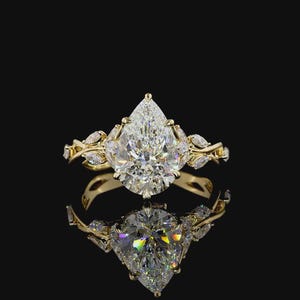 Puede incluir: Un anillo de oro con un gran diamante en forma de pera como pieza central. La banda presenta delicados detalles en forma de hojas y diamantes m&aacute;s peque&ntilde;os. El anillo se muestra sobre un fondo negro, con su reflejo visible debajo.