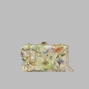 Op de afbeelding: Een rechthoekige clutch met een crèmekleurige basis en gedetailleerd bloemenborduursel in oranje, groene en blauwe tinten. De tas is versierd met doorzichtige lovertjes en kralen, afgewerkt met een goudkleurig metalen frame en een gouden kettinghengsel.