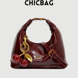 Peut inclure: Un sac à main en cuir verni bordeaux avec une poignée et une bandoulière dorées. Le sac est orné d'un charm en forme de cerise avec des cerises rouges et des feuilles dorées. Le texte "CHICBAG" est en haut.
