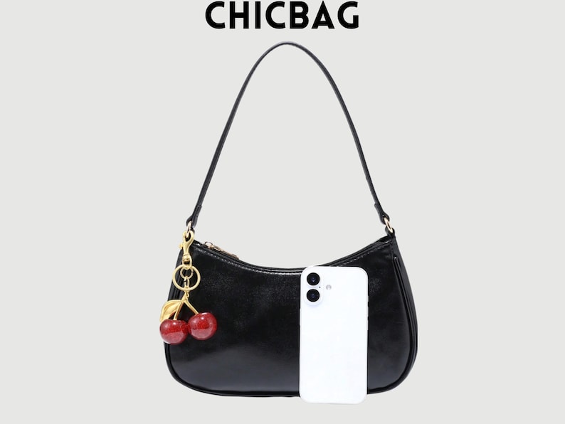 May include: Black shoulder bag with a gold zipper and a cherry charm. The bag has a curved shape and a long strap. The text "CHICBAG" is at the top. A white smartphone is next to the bag.