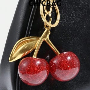 May include: A gold-toned metal keychain with a cherry design. The cherries are red with glitter, and the stem and leaf are gold. The keychain is attached to a black leather bag. The word "CHICBAG" is at the top.