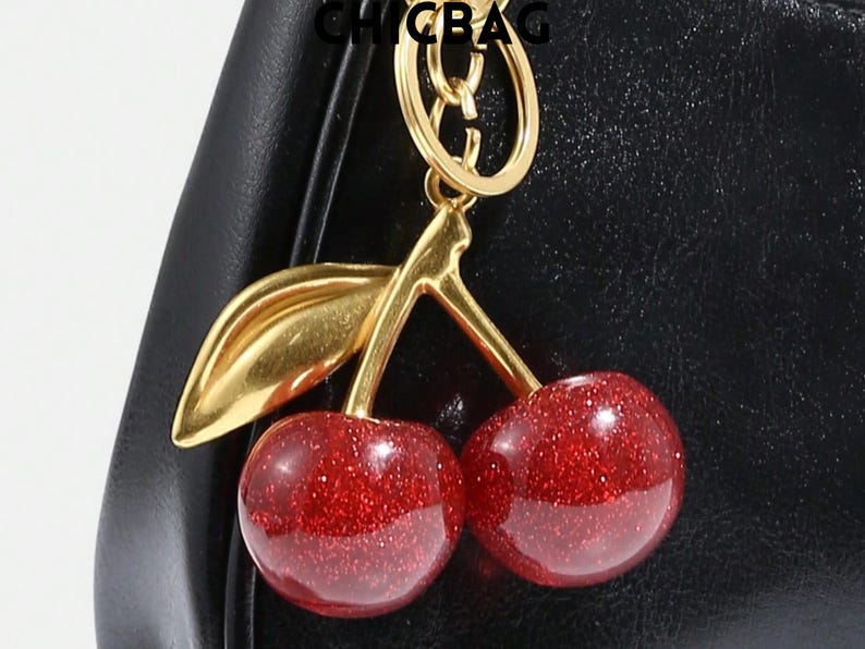 May include: A gold-toned metal keychain with a cherry design. The cherries are red with glitter, and the stem and leaf are gold. The keychain is attached to a black leather bag. The word "CHICBAG" is at the top.
