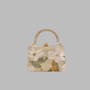 Op de afbeelding: Een rechthoekige handtas in een lichtbeige tint met witte lovertjes bloemmotieven en groene lovertjes bladeren. De tas is voorzien van een goudkleurig metalen frame, een bijpassend goudkleurig kralenhandvat en een kleine goudkleurige sluiting.