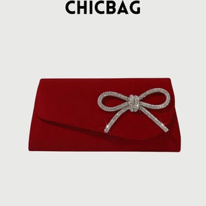 Puede incluir: Un clutch de terciopelo rojo con un lazo brillante de pedrería plateada. El lazo está centrado en la parte delantera de la bolsa. La palabra "CHICBAG" está impresa en la parte superior.