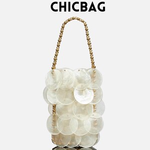 Puede incluir: Un bolso de mano blanco adornado con conchas, con un asa dorada con cuentas. El bolso está hecho de numerosas conchas redondas e iridiscentes, creando una textura única. El texto "CHICBAG" se muestra encima del bolso.