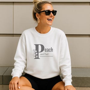 Könnte beinhalten: Weißes Sweatshirt mit Rundhalsausschnitt und dem Text "Peach and Pixel" in Grau. Das Design zeigt ein stilisiertes "P" mit floralen Akzenten. Das Model trägt schwarze Shorts und eine Sonnenbrille.